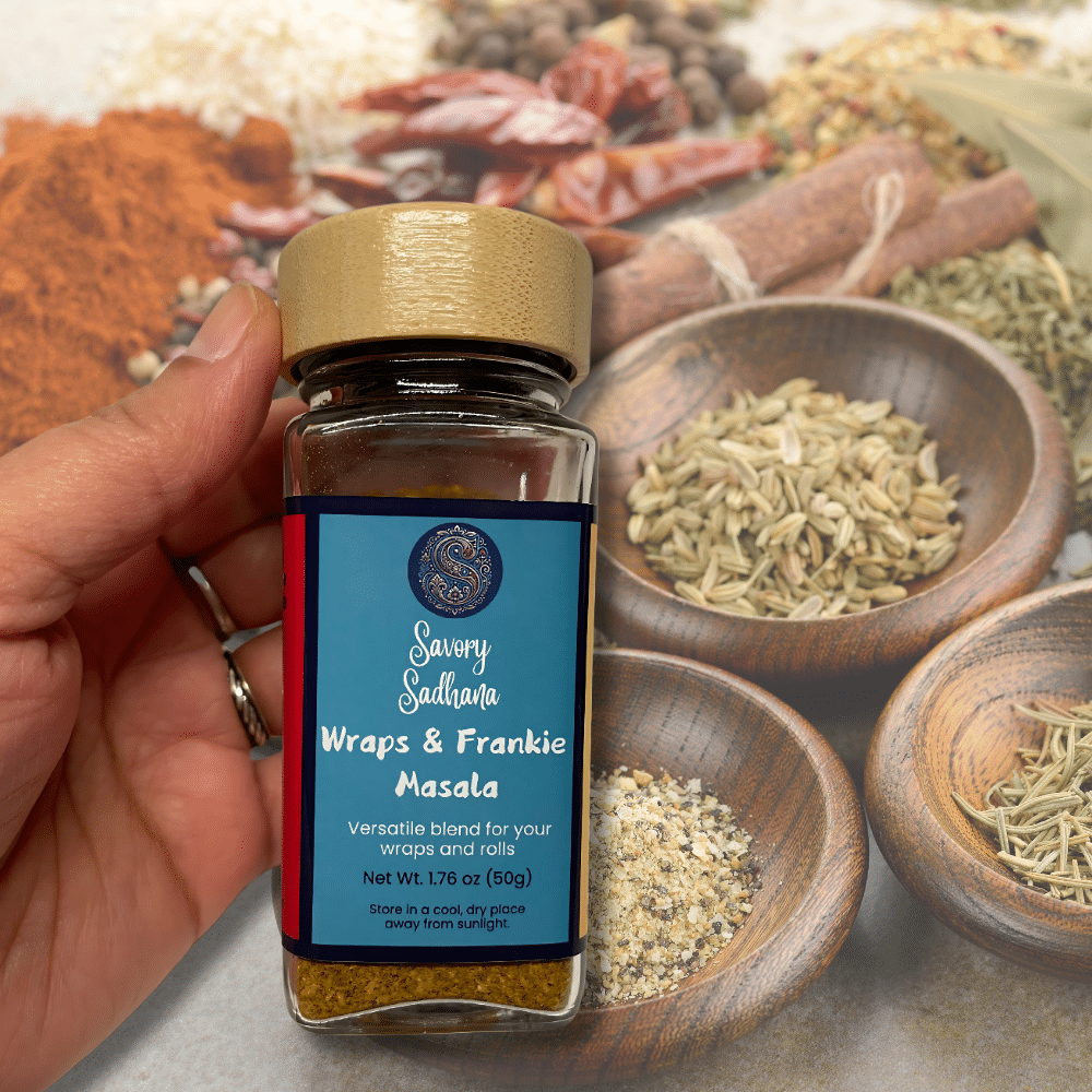 Hand holding a glass jar of Savory Sadhana Wraps & Frankie Masala with colorful whole spices in bowls blurred in the background.