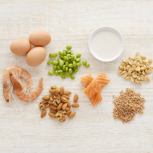 Top view of common food allergens including eggs, soybeans, milk, peanuts, shrimp, tree nuts, salmon, and wheat grains arranged on a light wooden surface.