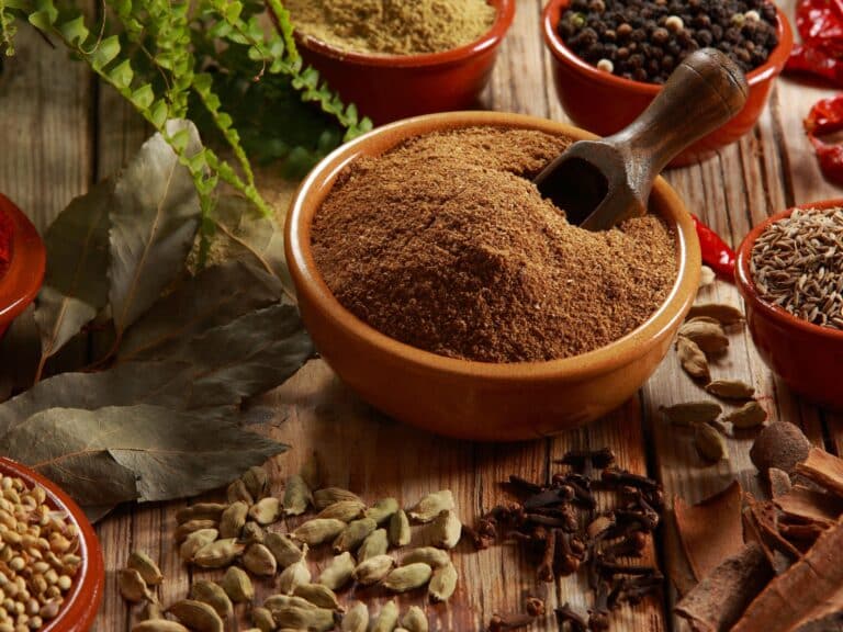 rich brown bowl of freshly ground heirloom garam masala surrounded by whole Indian spices like bay leaves, cardamom, cloves, cinnamon, black peppercorns, and cumin, arranged on a rustic wooden table.