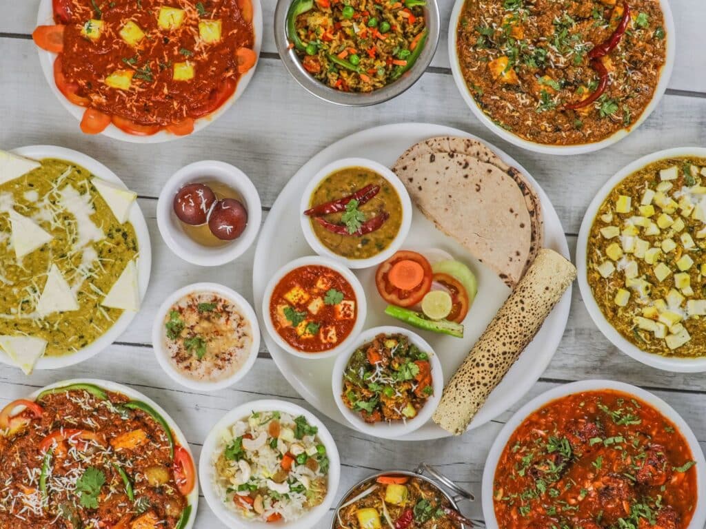 A vibrant spread of traditional Indian vegetarian dishes including paneer curries, dal, roti, rice, and pickles, all beautifully plated in white bowls on a white wooden table.