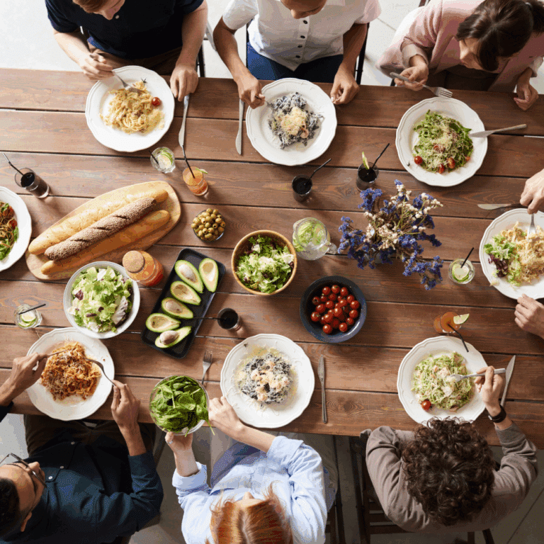 op-down view of a diverse group of people enjoying a colorful, healthy meal together at a rustic wooden table, featuring salads, pasta, avocado, bread, and fresh drinks—a celebration of mindful eating and community.